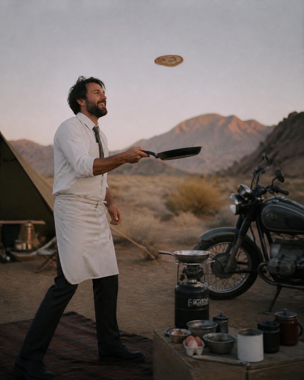 Man flipping pancakes beside a vintage motorcycle at a desert camp