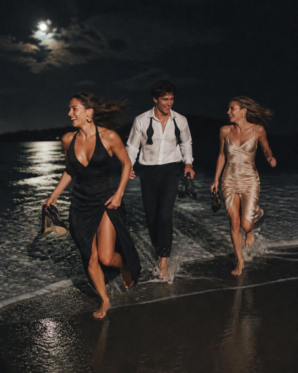 Friends in formal clothes running barefoot from the ocean at night