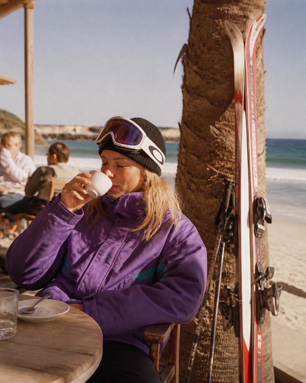 Woman in full ski gear drinking coffee at a beachside café