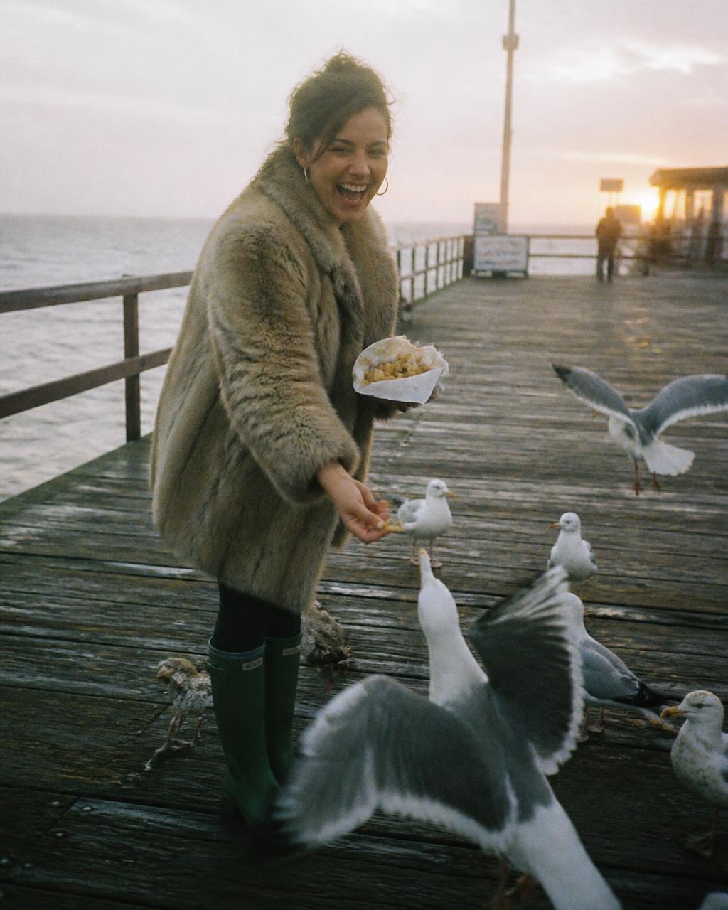 Woman in a fur coat feeding seagulls fries on a pier at sunset