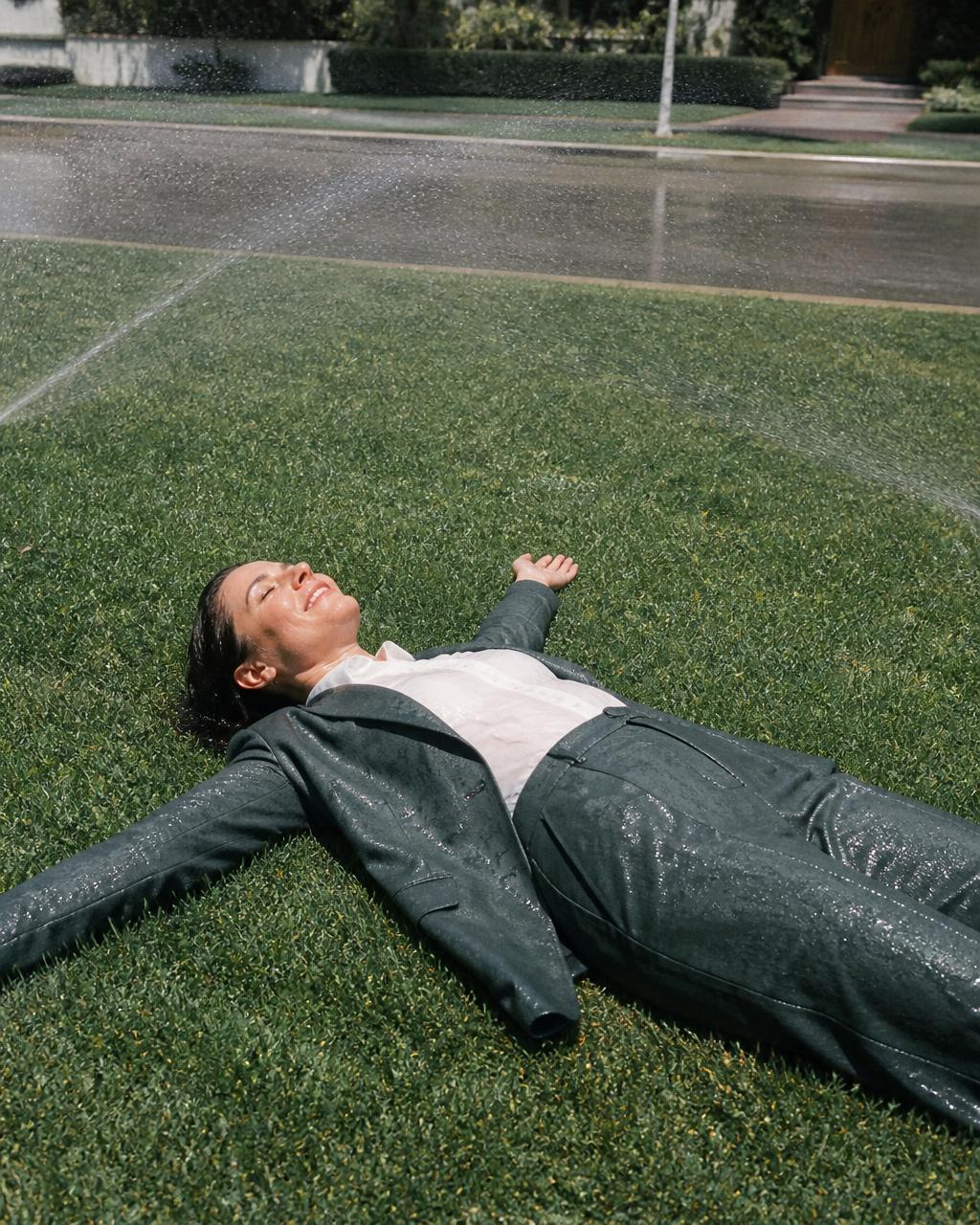 Man in a suit lying spread out on a suburban lawn in the sprinklers