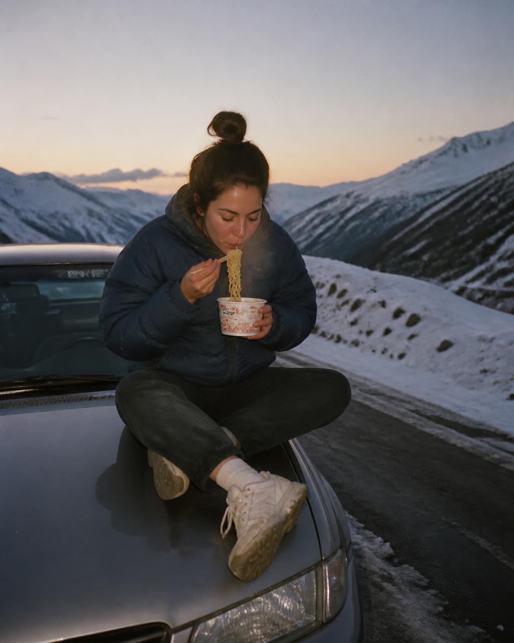 Woman in winter gear eating noodles on a car hood in the mountains