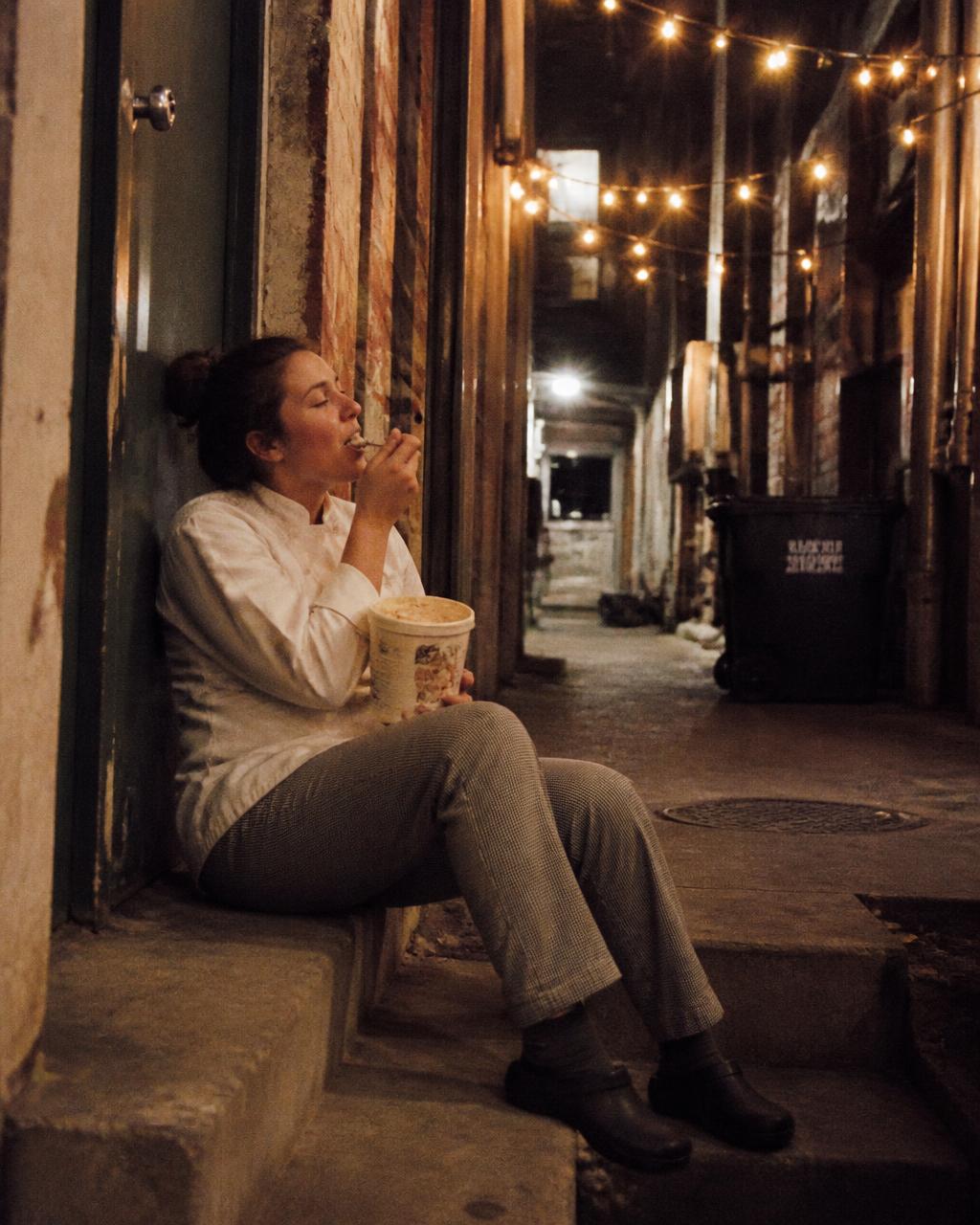 Chef eating ice cream from the tub on a back-alley step at midnight