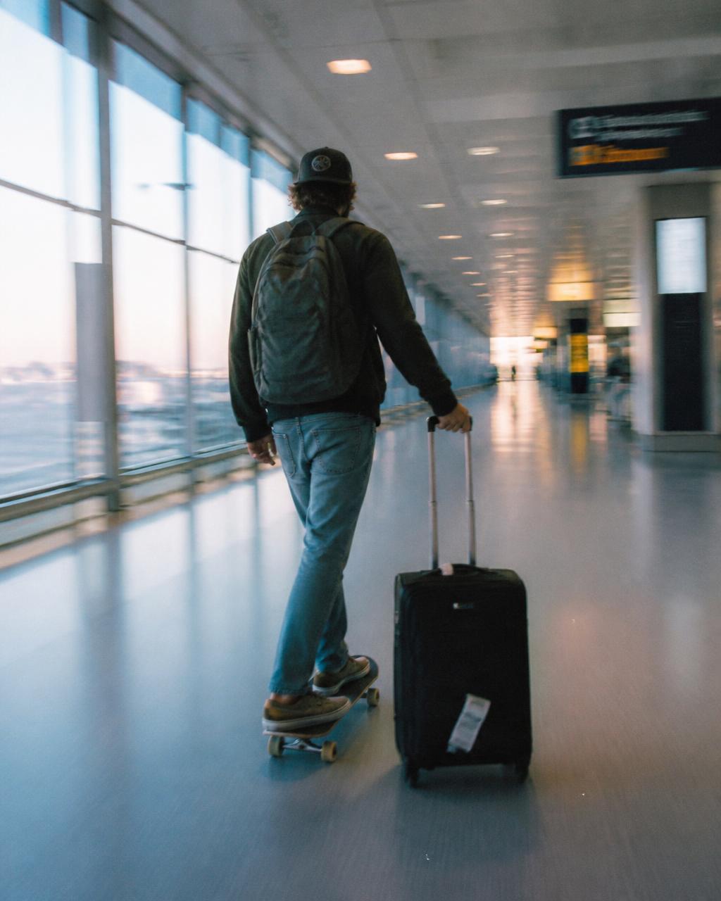 Man skateboarding through an airport terminal pulling a suitcase