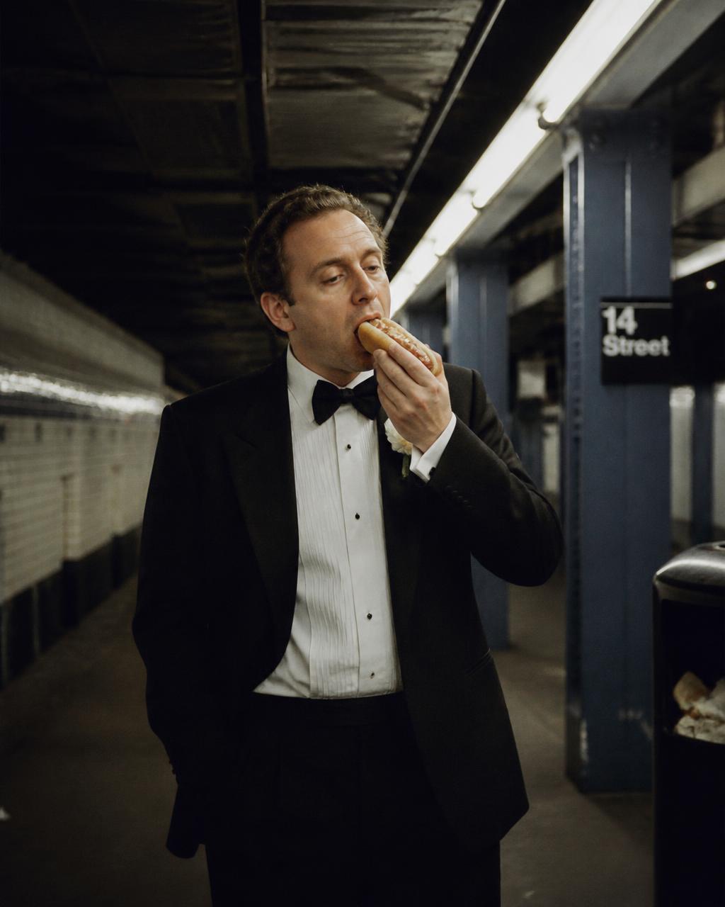 Man in a tuxedo eating a hot dog on a subway platform at 3am