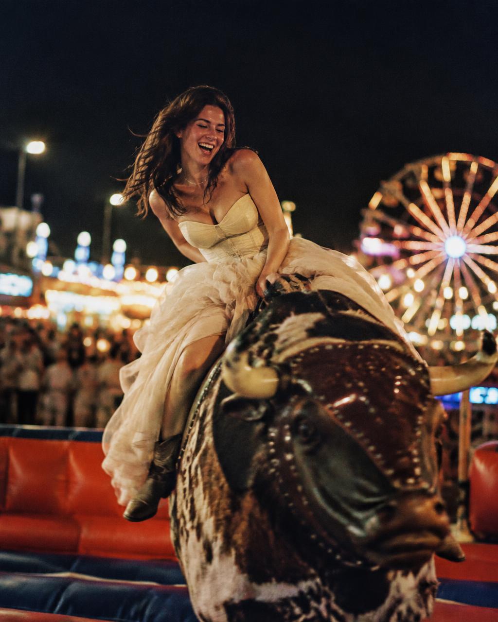 Woman in a ballgown riding a mechanical bull at a county fair