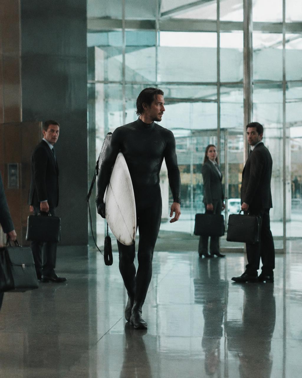Man in a wetsuit carrying a surfboard through an office lobby