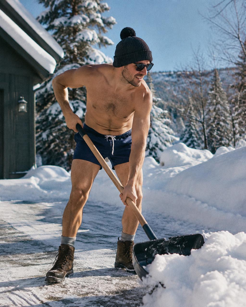 Man shoveling snow in swim trunks and a beanie